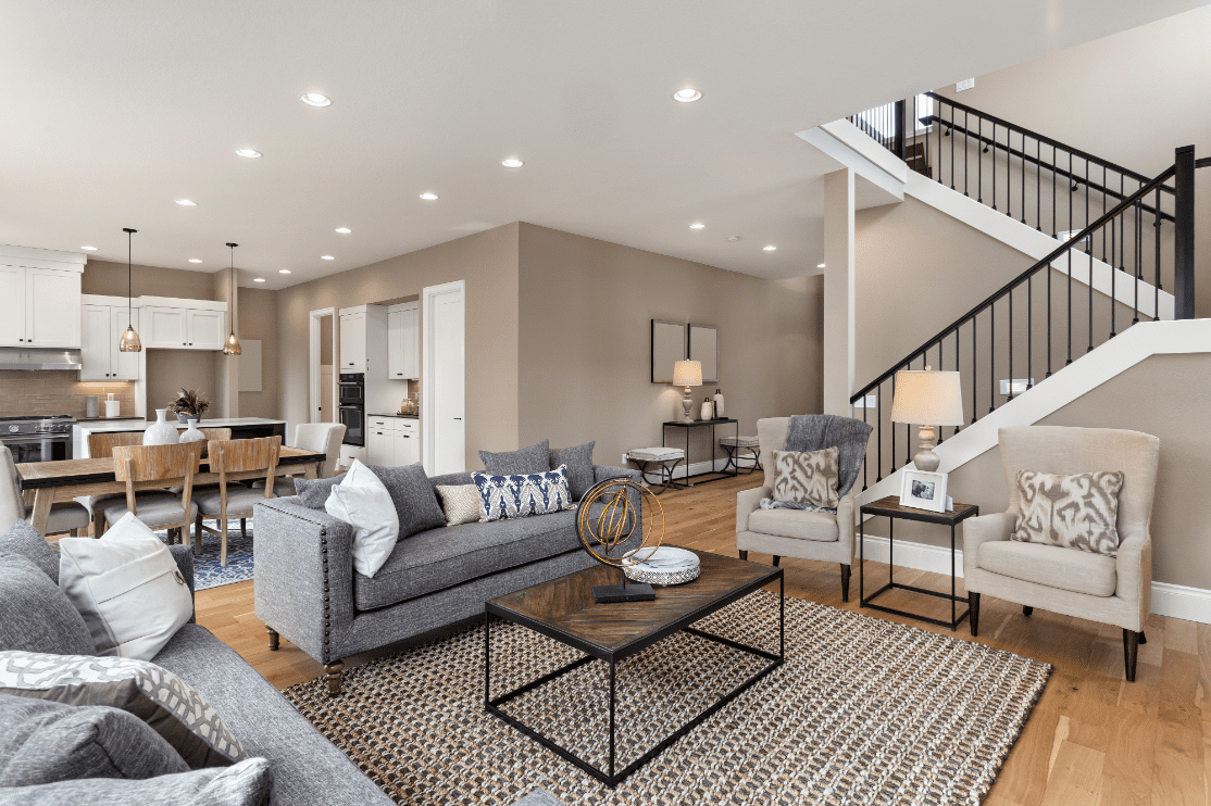 Spacious living room with hardwood floors, a gray sofa, armchairs, a coffee table, and a dining area. An open staircase leads to the upper level.