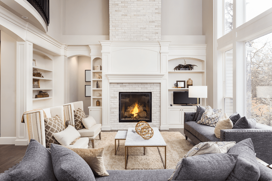 Modern living room with a lit fireplace, gray sofas, and white armchairs. A coffee table holds decorative items. Built-in shelves and a TV are on either side of the fireplace. Large windows let in natural light.