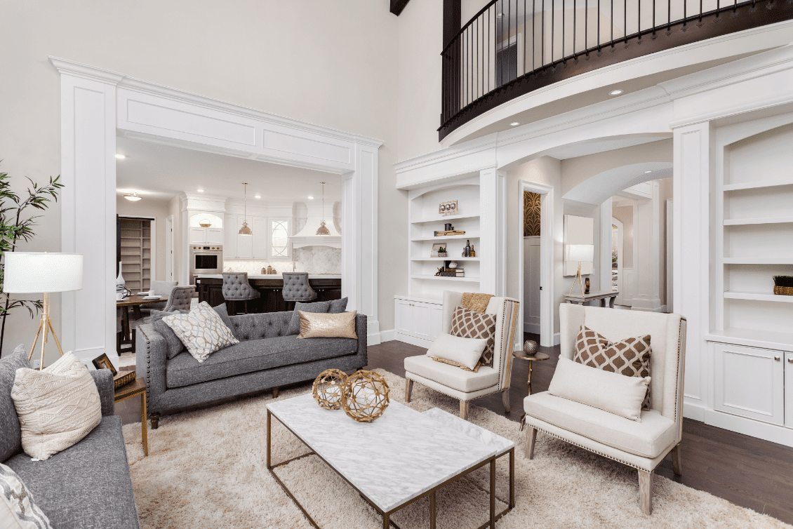 Elegant living room with a high ceiling, featuring gray sofas, plush chairs, a marble coffee table, and built-in shelves. An open archway leads to a modern kitchen in the background.