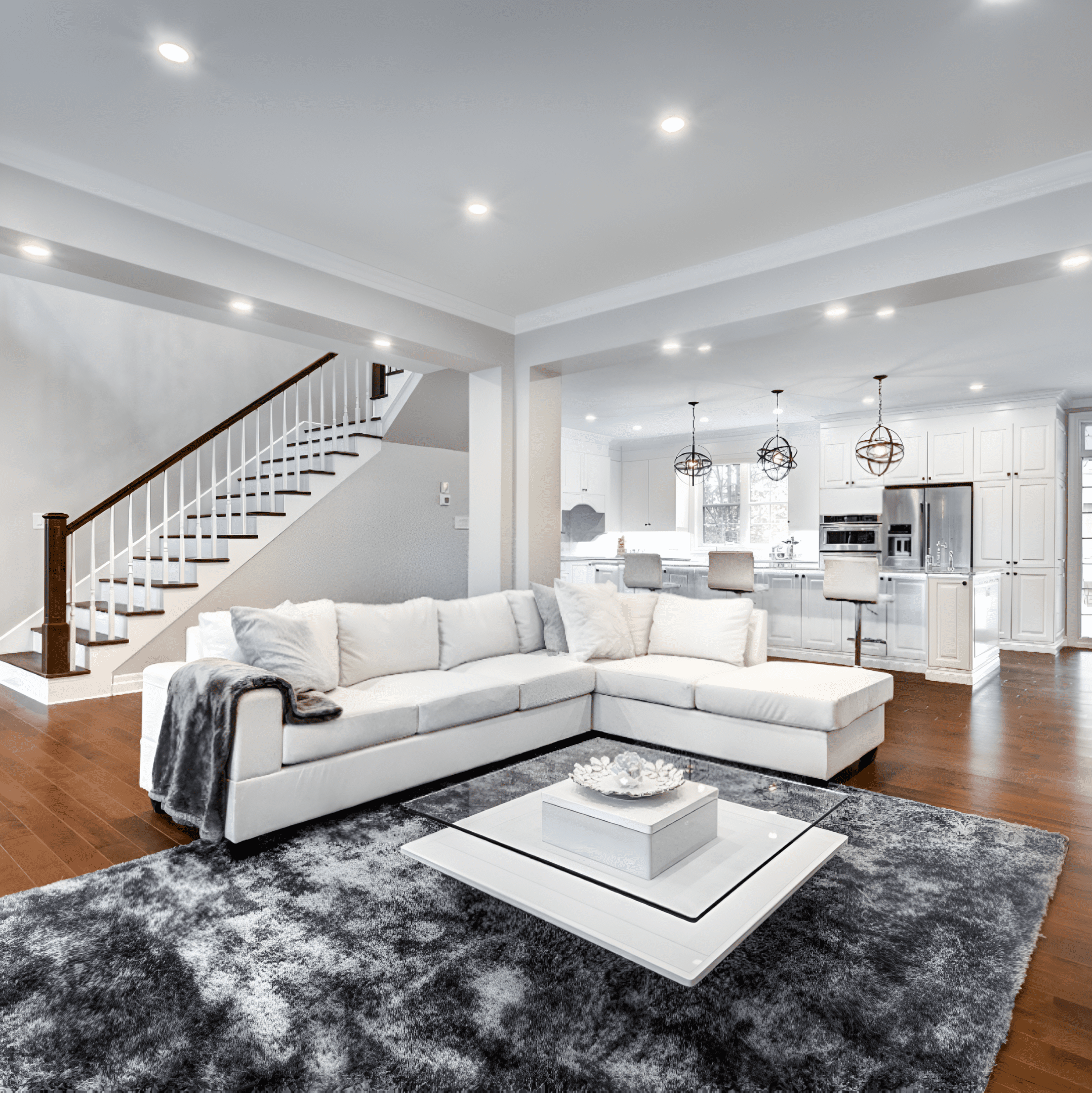 Modern open-concept living room with white sectional sofa, gray rug, and a central coffee table. Kitchen with white cabinets and island equipped with bar stools visible in the background.