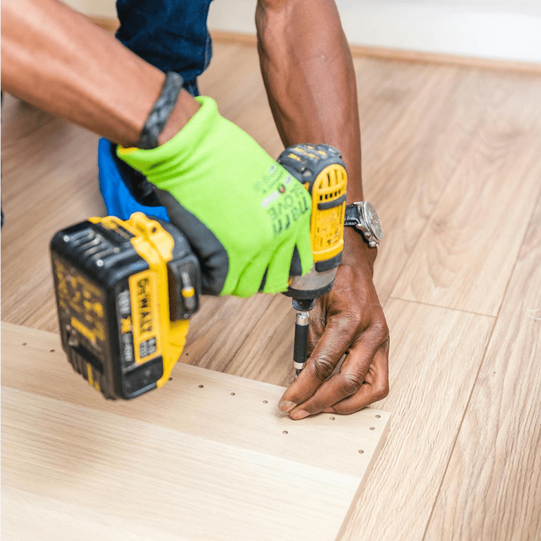 A person wearing a green glove uses a drill to insert screws into a light wooden panel on a hardwood floor.